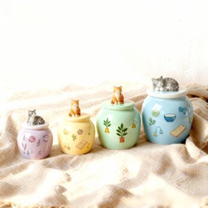 May include: Four ceramic jars in pastel shades, each with a cat figurine on top. The jars are of varying sizes and decorated with painted designs of plants, food, and other decorative elements. They are arranged on a beige fabric.