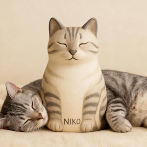 May include: A ceramic cat figurine with closed eyes and gray stripes, with the word "NIKO" printed on the front. A real cat is sleeping next to the figurine. The figurine is light beige and gray.