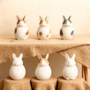 A group of six handmade ceramic rabbit urns in soft neutral colors, perfect memorial keepsakes for bunny ashes. Each personalized bunny urn features unique markings, cute shapes, and names, symbolizing warmth, comfort, and love for your pet rabbit. Ideal ceramic urn gift for rabbit lovers, memorial tribute for pets, handcrafted pottery urn with modern matte glaze for ashes, suitable for indoor home display, bunny memorial pottery for pet loss.