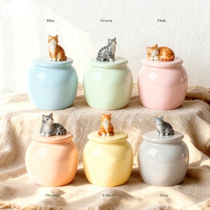 Peut inclure: Ensemble de six pots en céramique aux couleurs pastel : bleu, vert, rose, orange, jaune et gris. Chaque pot est surmonté d'un couvercle orné d'une figurine de chat. Les chats varient en couleur et en pose, ajoutant une touche ludique.
