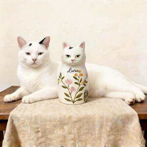 May include: A white ceramic cat figurine with the name "Luna" and floral designs. A real white cat with black markings is lying next to the figurine. The figurine is on a wooden table covered with a beige cloth.