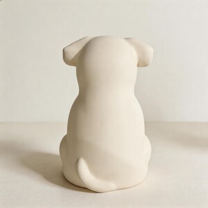 May include: A matte off-white ceramic figurine shaped like a dog, viewed from the back. The sculpture features a smooth, solid surface with rounded ears, a curved tail at the base, and a simple, clean silhouette representing a seated canine form.