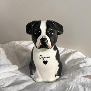 May include: A ceramic dog figurine featuring a black and white coat with floppy ears and a glossy finish. The chest area is white and displays the name Seymour in black script lettering above a small black heart icon centered on the front.