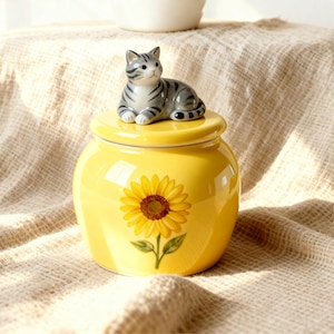 Könnte beinhalten: Ein gelbes Keramikgefäß mit Sonnenblumenmuster und einer grau-weißen Katzenfigur auf dem Deckel. Das Gefäß ist rund und hat eine glänzende Oberfläche. Die Sonnenblume ist mit grünen Blättern bemalt.