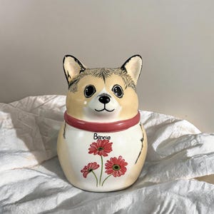 May include: A ceramic figurine shaped like a Corgi dog with tan and white fur, large black eyes, and a black nose. The dog wears a solid pink collar and has the name Bennie written above a cluster of three red daisy-like flowers on its chest.