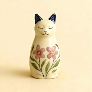 Op de afbeelding: Cr&egrave;mekleurig keramieken kattenbeeldje met gesloten ogen en een glimlachende uitdrukking. De kat heeft donkerblauwe oren en is versierd met twee roze bloemen en groene bladeren. Een decoratief item voor kattenliefhebbers.
