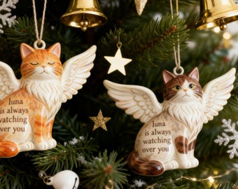 May include: Two ceramic cat ornaments with feathered wings, one orange tabby and one brown tabby. Each cat features the text 'luna is always watching over you' printed on its chest. The ornaments hang from strings against a green pine tree background.