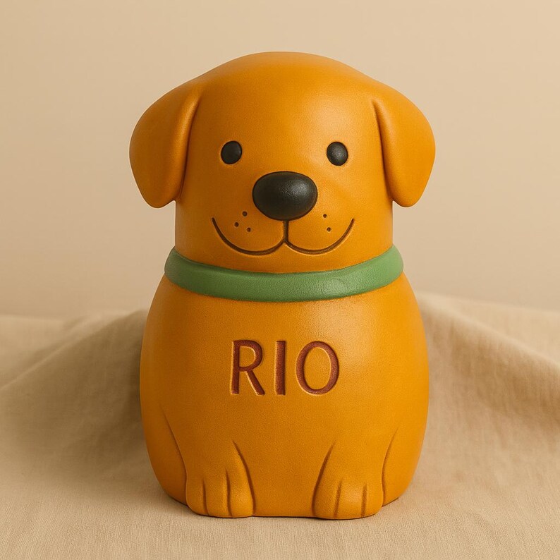 May include: A golden-yellow dog-shaped figurine with a smooth matte finish. The dog has floppy ears, a black nose, and a green collar. The name RIO is embossed in capital letters on the front of the dog's chest. The figure has a rounded body.