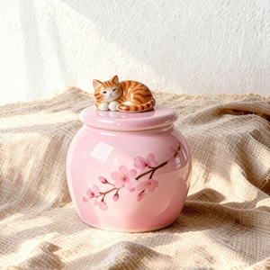 May include: A pink ceramic jar with a lid, adorned with a sleeping orange tabby cat figurine. The jar features a floral design of pink cherry blossoms. A decorative and functional item.