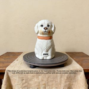 May include: A white ceramic dog figurine with an orange collar, sitting on a gray pottery wheel. The dog has detailed facial features and the name "Jake" is written on the front. The background is a neutral beige.