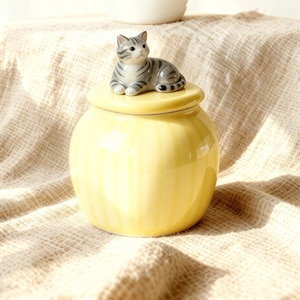 May include: A yellow ceramic jar with a lid, adorned with a small, grey and white striped cat figurine. The jar features vertical stripes and a rounded form. The background is a textured, off-white fabric.