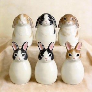 May include: Six ceramic bunny figurines in various colors, including white, brown, and black. The bunnies have detailed facial features and are arranged in two rows. The figurines are decorative and suitable for home decor.