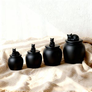 May include: Four black ceramic jars with cat figurines on the lids. The cats are in different poses, some sitting and one pair curled up. The jars are arranged on a textured, neutral-toned surface. The jars are in different sizes.