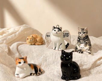 May include: Five small ceramic cat figurines in various poses and colors. One white cat has wings with the text Spud and It always watching over me. Others include a sleeping orange tabby, a sitting grey tabby, a calico cat, and a solid black cat.