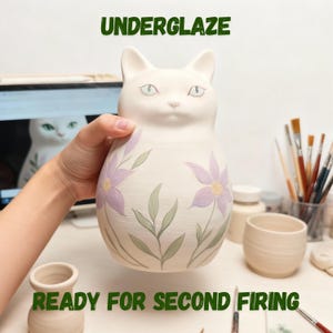 Op de afbeelding: Een witte keramische vaas in de vorm van een kat met paarse bloemen en groene bladeren. De teksten "UNDERGLAZE" en "READY FOR SECOND FIRING" zijn groen. De vaas wordt door een persoon vastgehouden, met kunstbenodigdheden en een computer op de achtergrond.
