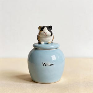May include: A light blue ceramic jar with a rounded body and a matching lid. The lid features a sculpted, hand-painted guinea pig figurine with black, white, and brown markings. The word Willow is printed in black, sans-serif letters on the jar.