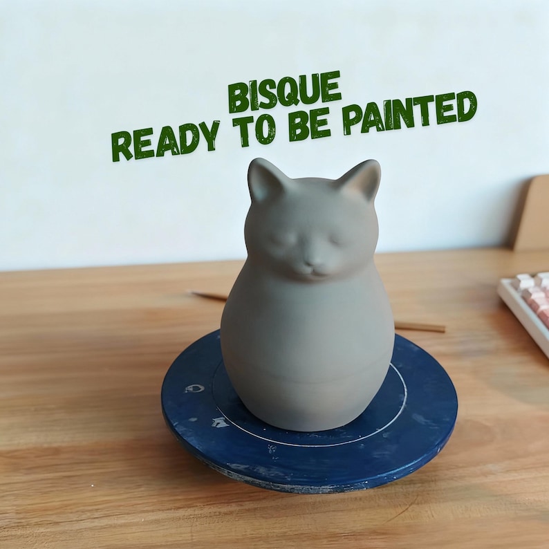Op de afbeelding: Een grijze keramische kattenfiguur, klaar om te worden beschilderd, staat op een blauw pottenbakkerswiel. De tekst "BISQUE READY TO BE PAINTED" is groen boven de kat gedrukt. De kat heeft gesloten ogen en puntige oren.