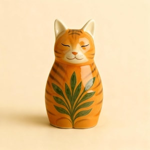 Op de afbeelding: Een keramieken kattenbeeldje met oranje en bruine strepen, gesloten ogen en een groen bladerontwerp op de borst. De kat heeft witte oren en een lachend gezicht, wat een grillig en decoratief stuk cre&euml;ert.