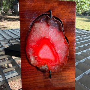 May include: A decorative wooden plaque featuring a vibrant, red and pink agate slice. The agate is mounted on a polished, reddish-brown wooden base. A dark metal rod runs through the agate. The piece is displayed outdoors.
