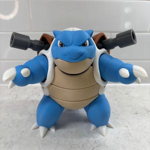 May include: A blue and tan Blastoise figurine with white accents and two gray cannons on its back. The figurine has a determined expression and white claws. The figure is standing on a light gray surface.