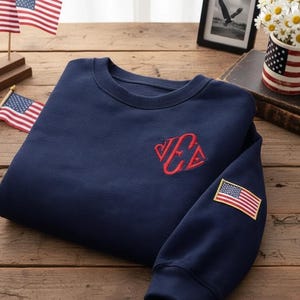 May include: A navy blue sweatshirt with a red embroidered monogram and an American flag patch on the sleeve. The sweatshirt is folded on a wooden surface, with small American flags and a mug with an American flag design in the background.