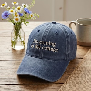 Puede incluir: Gorra de béisbol de mezclilla azul con el texto bordado "I'm coming to the cottage". La gorra está sobre una superficie de madera, con un jarrón de flores y una taza en el fondo.