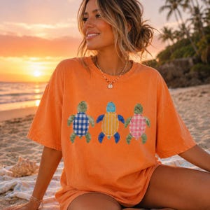 May include: An orange short-sleeve t-shirt featuring a graphic print of three sea turtles. The left turtle has a blue and white gingham shell, the center turtle has a yellow and white striped shell, and the right turtle has a pink and white plaid shell.