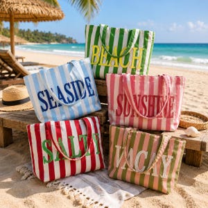 Op de afbeelding: Vijf rechthoekige canvas draagtassen met verticale strepen en geborduurde tekst. De tassen tonen de woorden BEACH in geel, SEASIDE in blauw, SUNSHINE in rood, SALTY in groen en VACAY in wit, elk weergegeven op een bijpassende gestreepte achtergrond.