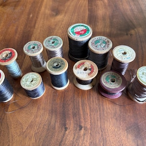 May include: A collection of twelve vintage wooden spools of sewing thread in various colors including black, grey, and purple. Each spool features a circular paper label on top with printed brand names such as Rice's, Gudebrod, and Thairwall & Co.