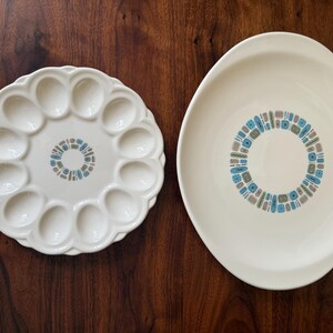 May include: A round white ceramic egg plate with twelve indentations and a matching oval serving platter. Both pieces feature a central circular pattern composed of small blue, green, and grey geometric shapes on a smooth off-white surface.