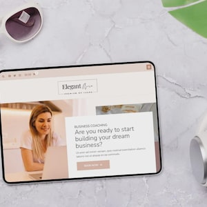 Op de afbeelding: Een digitale tablet toont een website met de tekst "Elegant Aria" en "Premium of Icons". Het scherm toont een vrouw die op een laptop werkt. Witte zonnebril en koptelefoon zijn ook zichtbaar.
