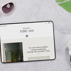 Puede incluir: Una tableta digital que muestra el sitio web "GOLDEN THREADS", con el texto "Are you ready to build a content marketing strategy that actually works?". Tambi&eacute;n se ven gafas de sol y auriculares blancos.