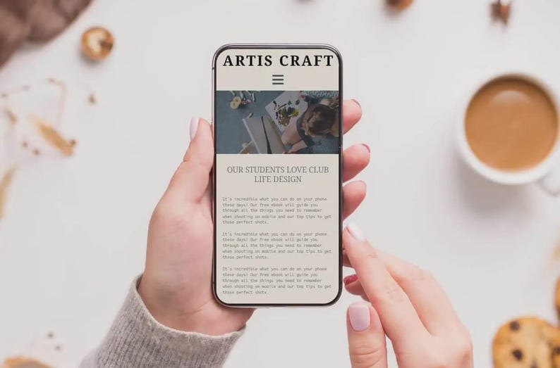 Puede incluir: Un smartphone muestra el sitio web "ARTIS CRAFT" y "OUR STUDENTS LOVE CLUB LIFE DESIGN". El tel&eacute;fono lo sostiene una persona. Una taza de caf&eacute; y galletas est&aacute;n en el fondo.