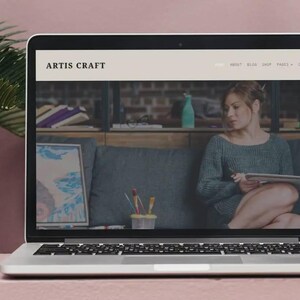 Puede incluir: Un ordenador port&aacute;til muestra el sitio web "ARTIS CRAFT". El sitio web presenta a una mujer sosteniendo una tableta. Se ven una obra de arte enmarcada y materiales de arte. El port&aacute;til est&aacute; sobre una superficie rosa con una hoja de palma.