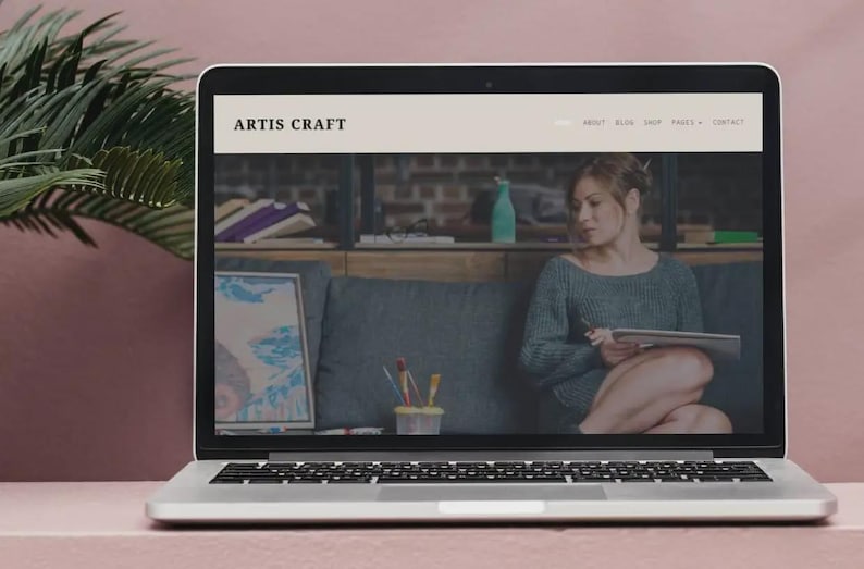 Puede incluir: Un ordenador port&aacute;til muestra el sitio web "ARTIS CRAFT". El sitio web presenta a una mujer sosteniendo una tableta. Se ven una obra de arte enmarcada y materiales de arte. El port&aacute;til est&aacute; sobre una superficie rosa con una hoja de palma.