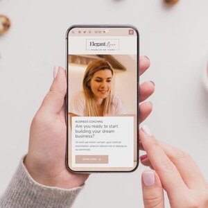Op de afbeelding: Een smartphone toont de website voor "Elegant Aura" business coaching. Het scherm toont een vrouw en de tekst "Ben je klaar om je droombedrijf te starten?" De telefoon wordt door een persoon vastgehouden, met een kop koffie en koekjes.