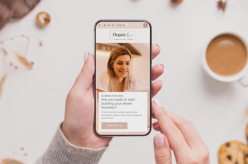Op de afbeelding: Een smartphone toont de website voor "Elegant Aura" business coaching. Het scherm toont een vrouw en de tekst "Ben je klaar om je droombedrijf te starten?" De telefoon wordt door een persoon vastgehouden, met een kop koffie en koekjes.