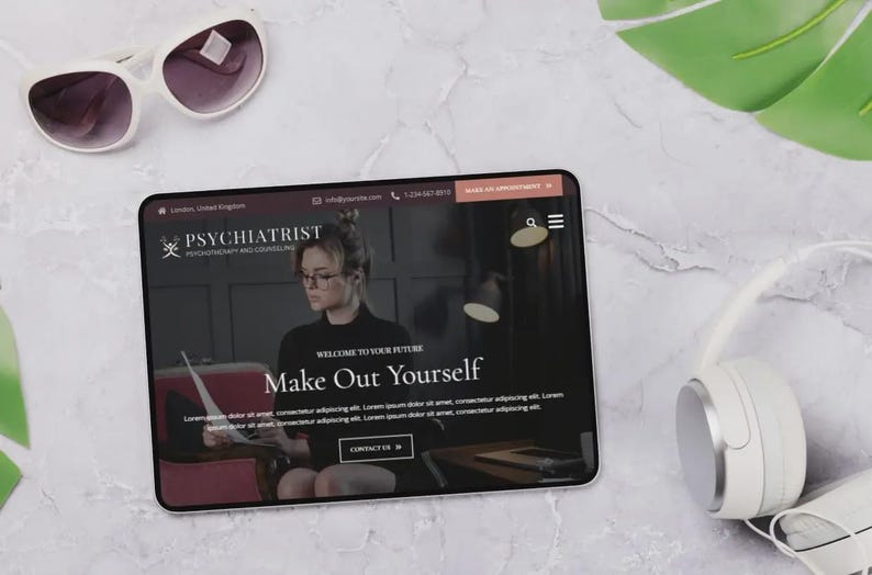 May include: A tablet displaying a website for a psychiatrist, with the text "Make Out Yourself." White sunglasses and headphones are next to the tablet. The website features a woman wearing glasses. The background is a marble surface.