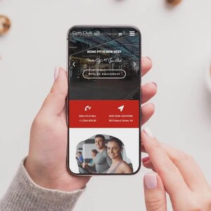May include: A smartphone displaying a website for Gym Club, with the text "BEING FIT IS NEW SEXY". The phone is held by a person, with a cup of coffee and cookies in the background. The website includes a call to action to make an appointment.