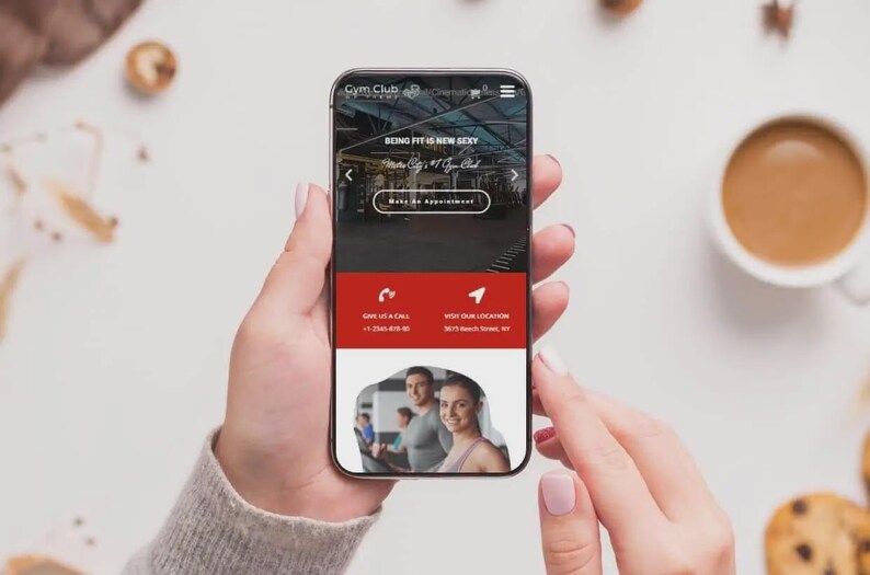 May include: A smartphone displaying a website for Gym Club, with the text "BEING FIT IS NEW SEXY". The phone is held by a person, with a cup of coffee and cookies in the background. The website includes a call to action to make an appointment.