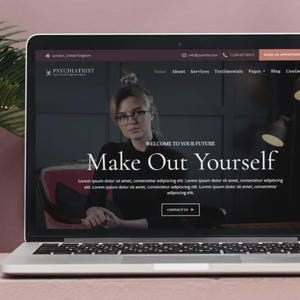 May include: A laptop displays a website for a psychiatrist, featuring a woman in glasses. The site's headline reads "Make Out Yourself." The screen shows a dark background with text and a contact button. The laptop is open on a pink surface.