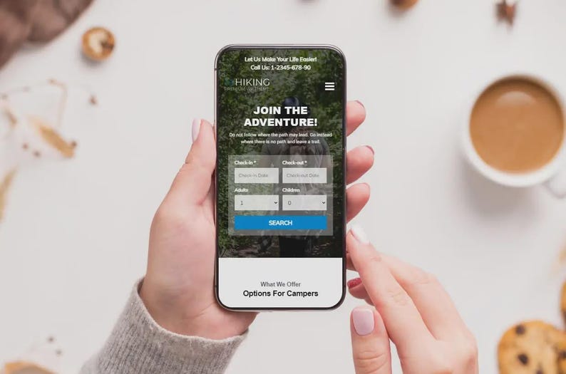 May include: A smartphone screen shows a hiking website with the text "Join the Adventure!". The phone displays a search form and a phone number. The phone is held by a person, with a cup of coffee and biscuits in the background.