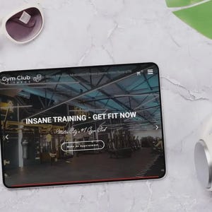 May include: A tablet displays a gym website with the text "INSANE TRAINING - GET FIT NOW." White sunglasses and headphones are next to the tablet. The background is a marble-like surface with a green leaf.