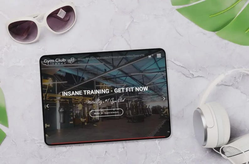 May include: A tablet displays a gym website with the text "INSANE TRAINING - GET FIT NOW." White sunglasses and headphones are next to the tablet. The background is a marble-like surface with a green leaf.