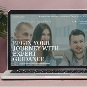 May include: A silver laptop shows a website with the text "Business Coach" and the tagline "Begin Your Journey With Expert Guidance." The screen displays three people. The laptop is on a pink surface with a plant in the background.