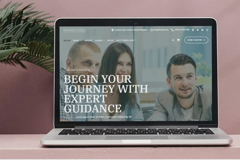 May include: A silver laptop shows a website with the text "Business Coach" and the tagline "Begin Your Journey With Expert Guidance." The screen displays three people. The laptop is on a pink surface with a plant in the background.