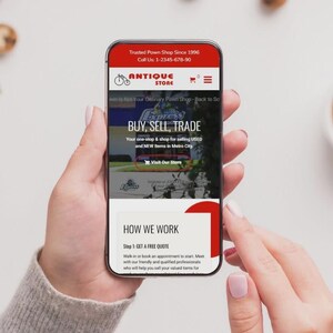 May include: A smartphone displaying an antique store website, held in a person's hands. The website has a red and white design with the text "BUY, SELL, TRADE" and "ANTIQUE STORE". A cup of coffee and biscuits are in the background.