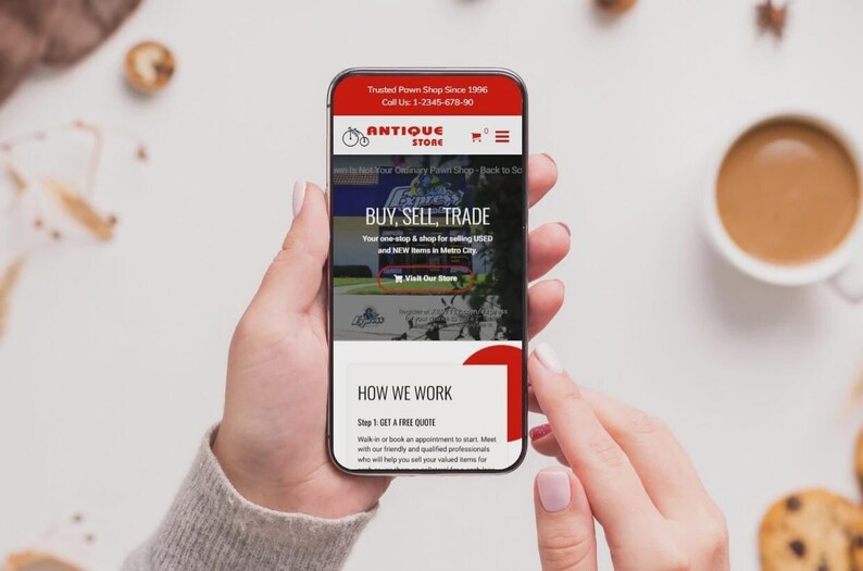 May include: A smartphone displaying an antique store website, held in a person's hands. The website has a red and white design with the text "BUY, SELL, TRADE" and "ANTIQUE STORE". A cup of coffee and biscuits are in the background.