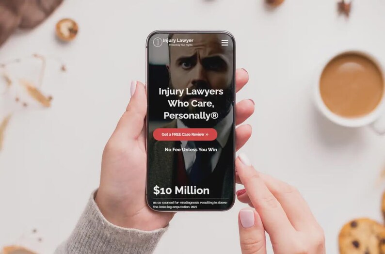May include: A smartphone displays a website for Injury Lawyers. The screen features the text "Injury Lawyers Who Care, Personally&reg;" with a red button that says "Get a FREE Case Review." The phone is held by a person, with a cup of coffee and cookies in the background.