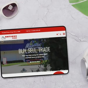 May include: A tablet showing a website with the words "BUY, SELL, TRADE" and "ANTIQUE STORE". White sunglasses and headphones are placed beside the tablet. The website also says "Trusted Pawn Shop Since 1996".